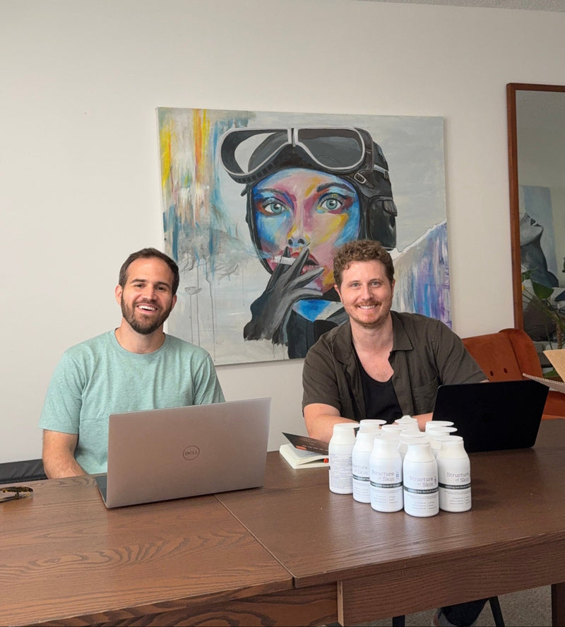 Two men sitting at a desk with a laptop and bottles, with a colorful painting in the background.